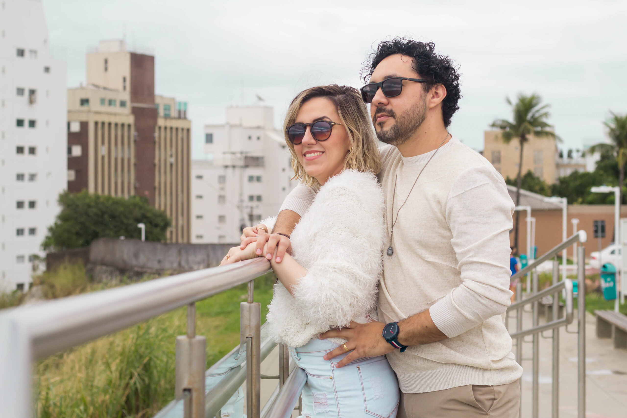 Dra. Natasha Veloso e Dr. Yuri Sena de óculos escuros, lado a lado, contemplando o mar da Praia da Enseada no Guarujá, em um momento íntimo de conexão