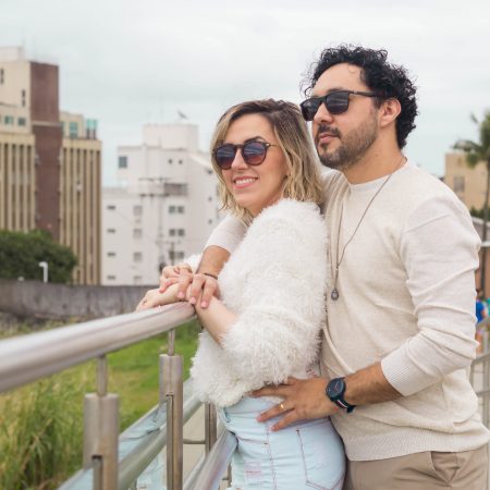 Dra. Natasha Veloso e Dr. Yuri Sena de óculos escuros, lado a lado, contemplando o mar da Praia da Enseada no Guarujá, em um momento íntimo de conexão
